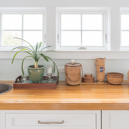 Kitchen counter styling with butcher block top, white cabinets, woven storage baskets, potted plant, and vintage serving tray under windows